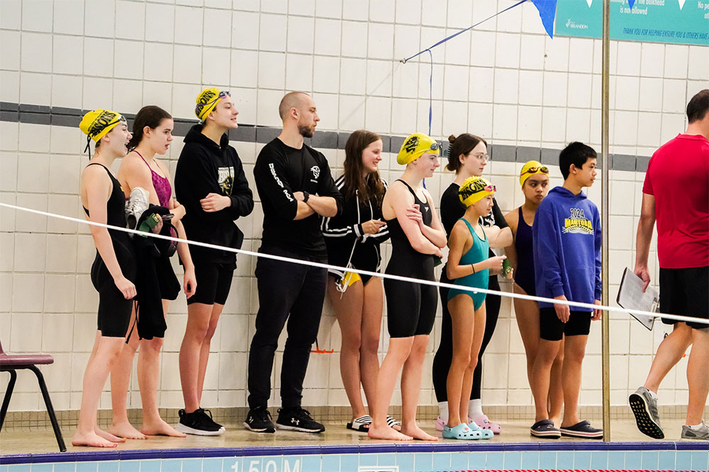 Swimmers of various ages stand against the wall near the pool