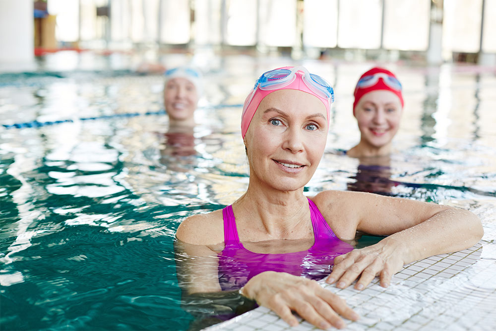 Adult female swimmers wearing swimming caps, treading water in pool. One woman has her arm resting on the poolside, smiling happily